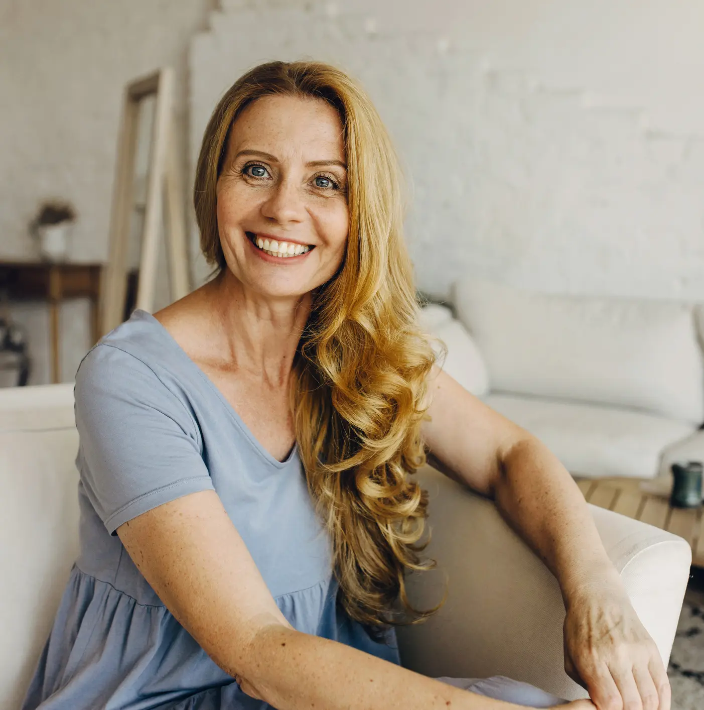 ttractive adult woman sitting on coach in living-room with loft design, having beautiful, clean and combed wavy hair, big blue eyes matching with color of her dress. Natural beauty concept Facelift in St. Louis, MO